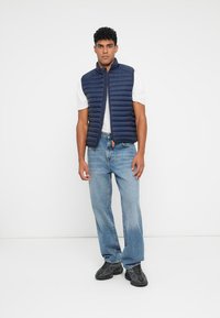 Marineblå polstret vest med lynlås foran, let design, uden ærmer, i kombination med en hvid t-shirt og blå jeans, stående på en neutral baggrund.