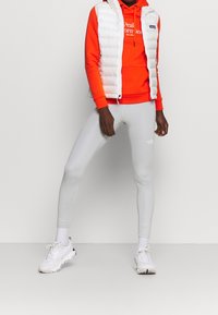Veste matelassée blanche portée sur un sweat à capuche orange, associée à des leggings gris et des baskets blanches. Textures lisses et coupe dynamique dans l'ensemble.