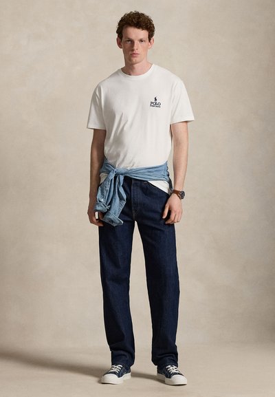 Junger Mann trägt ein weißes Polo Ralph Lauren T-Shirt, dunkelblaue Jeans, blaue Sneakers und ein Jeanshemd, das um seine Taille gebunden ist.