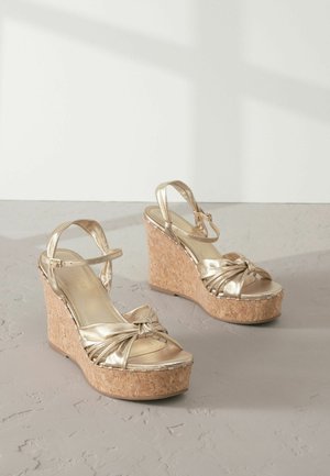 Gold wedge sandals with a cork platform, featuring a twisted knot detail on the front and adjustable ankle straps.