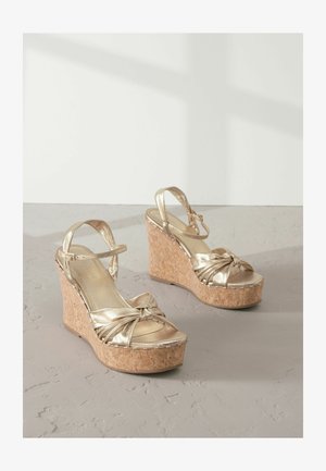 Zlatne wedge sandale s plutenom platformom, koje imaju zakrivljen čvor na prednjem dijelu i podesive naramenice za gležnjeve.
