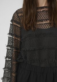 Sieviete valkā melnu caurspīdīgu mežģīņu topu ar ģeometriskiem rakstiem un frēgām horizontālajās svītrās, kas kombinēts ar melniem savelktiem auduma elementiem apakšā.