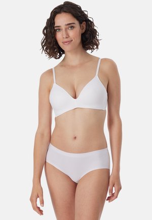 Femme aux cheveux bouclés foncés portant un soutien-gorge blanc sans couture et un slip blanc assorti sans couture, debout devant un fond clair uni.