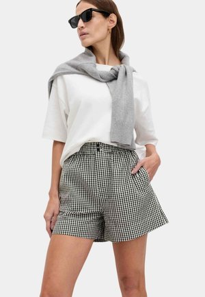 Kvinde iført sort-hvide gingham-shorts, oversize hvid T-shirt, grå sweater hængende over skuldrene og sorte solbriller, hånd i lommen.