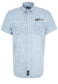 Valkoinen lyhythihainen farkkupaita, jossa on napitus ja kaksi napillista rintataskua, sekä musta painatus vasemmalla ylhäällä.