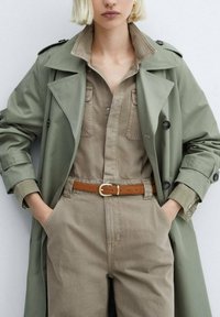 Manteau trench vert olive sur une combinaison beige, avec un col, des poches et une ceinture marron. Texture lisse avec des accents de boutons.