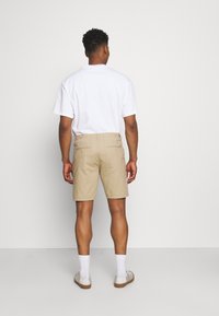 Khaki szorty o luźnym kroju, z elastycznym paskiem, tylnymi kieszeniami i gładką fakturą. Noszone z białą koszulką i butami.