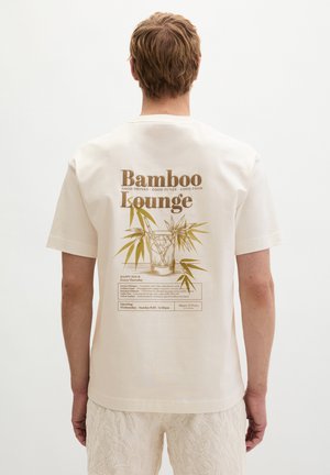 Man draagt een wit T-shirt met de tekst 'Bamboo Lounge' en een illustratie van een bamboeplant op de achterkant, gecombineerd met licht gestructureerde korte broek.