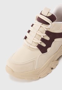 Chunky beige sneaker with thick sole, cream laces, and dark brown suede accents on sides and tongue.