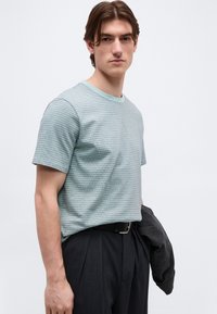 Νεαρός άνδρας με μεσαίου μήκους μαλλιά που φοράει ανοιχτό μπλε ριγωτό κοντομάνικο πουκάμισο και σκούρο παντελόνι, κρατώντας ένα σκούρο σακάκι στο ένα χέρι.