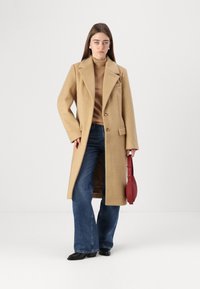 Lungo cappotto di lana color cammello con risvolti, due bottoni e una tasca laterale. Indossato sopra un top marrone e jeans a gamba larga blu, mentre tiene una borsa rossa.