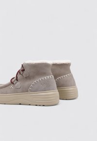 Botines de ante beige con un suave forro de borreguito blanco, que cuentan con cordones marrones y costuras decorativas en la parte superior y en la entresuela.
