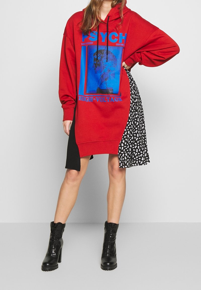 Femme portant un sweat à capuche rouge surdimensionné avec un texte graphique bleu « PSYCH HIGH-VOLTAGE », un panneau de jupe fleurie noir et blanc, et des bottes à talons noires.