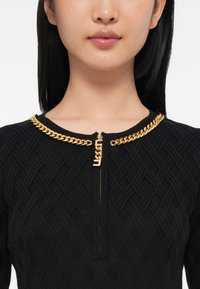Gros plan sur une personne portant un haut texturé noir avec un détail en chaîne dorée autour du col et une tirette en or.