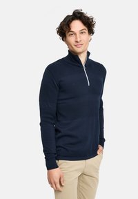 Marineblaue gestrickte Sweatjacke mit einem halben Reißverschlusskragen, strukturierten Mustern, gerippten Bündchen und Saum. Trägt man über hellen khakifarbenen Hosen.