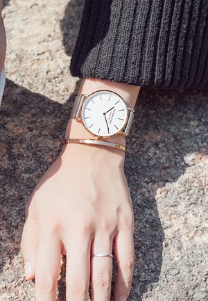 Reloj con correa de malla plateada, cara blanca y diseño minimalista. Acompañado de una pulsera dorada y un anillo plateado delgado en una mano.