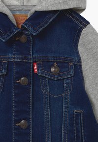 Dark blue denim jacket with brown stitching, metal buttons, front pockets, gray fabric sleeves, and attached gray hood.