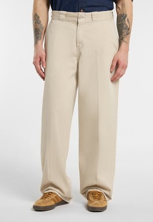 Pantaloni beige ampi con bottone e zip frontali, indossati con sneakers marroni e una camicia scura, che mostrano avambracci tatuati.