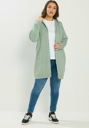 Frau trägt einen hellgrünen Hoodie mit Reißverschluss, weißes T-Shirt, blaue Jeans und schwarze Turnschuhe, steht vor einem schlichten weißen Hintergrund.