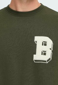 Nærbilde av en olivengrønn crewneck sweatshirt med en teksturert hvit bokstav "B" applikert på brystet.