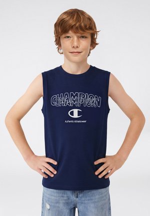 Ragazzo dai capelli rossi che indossa una maglietta smanicata Champion blu navy e jeans azzurri, in piedi con le mani sui fianchi contro uno sfondo semplice.