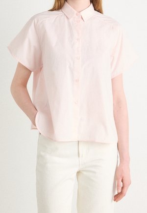 Personne portant une chemise à manches courtes rose clair boutonnée et un pantalon blanc cassé, debout avec une main dans la poche devant un fond uni.