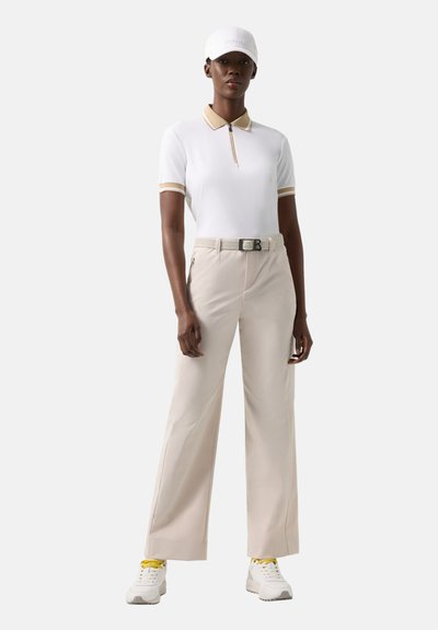 Femme debout portant un polo blanc avec des bordures beiges, un pantalon large beige ceinturé, des baskets blanches à lacets jaunes, et une casquette blanche.