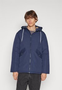 Manteau matelassé bleu marine avec capuche, cordons de serrage, fermeture éclair à l'avant et deux poches latérales avec rabats. Doublure intérieure offrant une chaleur supplémentaire.