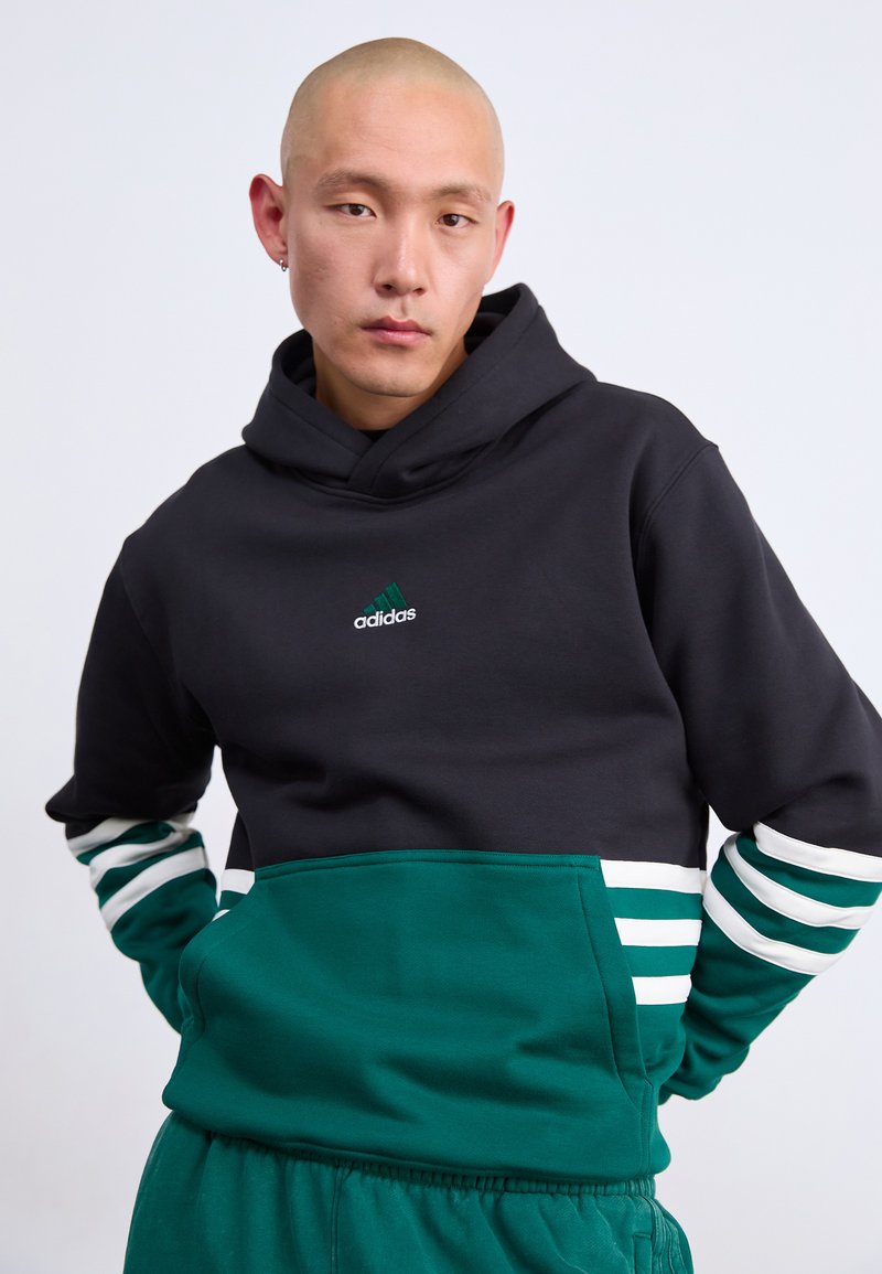 Joven calvo con sudadera Adidas negra y verde con rayas blancas, de pie con las manos detrás de la espalda frente a un fondo plain.