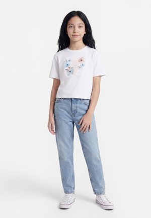 Dziewczyna z długimi ciemnymi włosami, ubrana w białą koszulkę z kwiatowym nadrukiem motyli, jasnoniebieskie dżinsy i białe trampki, stojąca na białym tle.