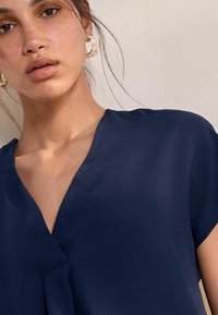 Blusa blu navy con scollatura a V e maniche corte, realizzata in un tessuto liscio e lucido, con una vestibilità comoda. I dettagli accentuati includono una piega sottile.