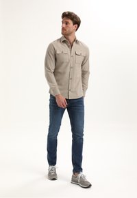 Camicia button-up beige chiaro con due tasche sul petto, abbinata a jeans blu e sneakers grigie. Design semplice con un tessuto testurizzato.