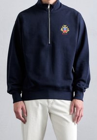 Felpa zip con collo alto di colore blu navy, con polsini ed orlo elasticizzati. Presenta un logo ricamato in giallo e rosso sul petto.