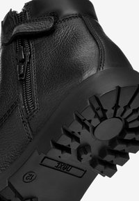 Zwarte leren enkellaars met een zijrits en klittenbandsluiting. Dikke rubberen zool met een gestructureerd loopprofiel voor grip.