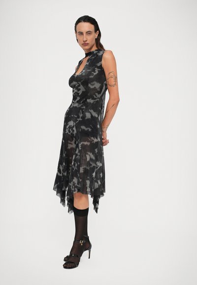 Vrouw met teruggekamd haar, gekleed in een mouwloze zwart-grijze camouflage jurk met een asymmetrische zoom, doorschijnende zwarte sokken en zwarte hoge hakken.