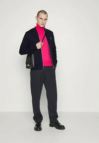 Tenue pour homme comprenant une veste bomber bleu marine, un col roulé rose vif, un pantalon sombre et des chaussures noires. Comprend un sac à bandoulière noir.