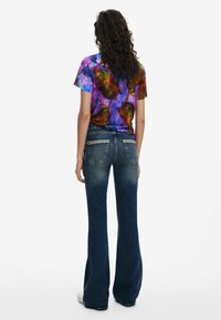 Camicia a maniche corte multicolore con motivo astratto, abbinata a jeans a zampa d'elefante blu scuro con due tasche posteriori e accenti sbiaditi.