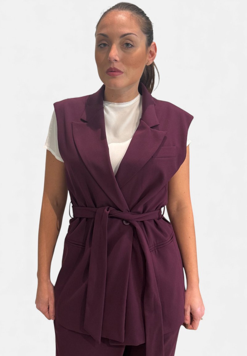 Blazer bordeaux senza maniche con vita cinturata, colletto e tasche frontali. Indossato sopra una maglietta bianca, con una texture liscia.