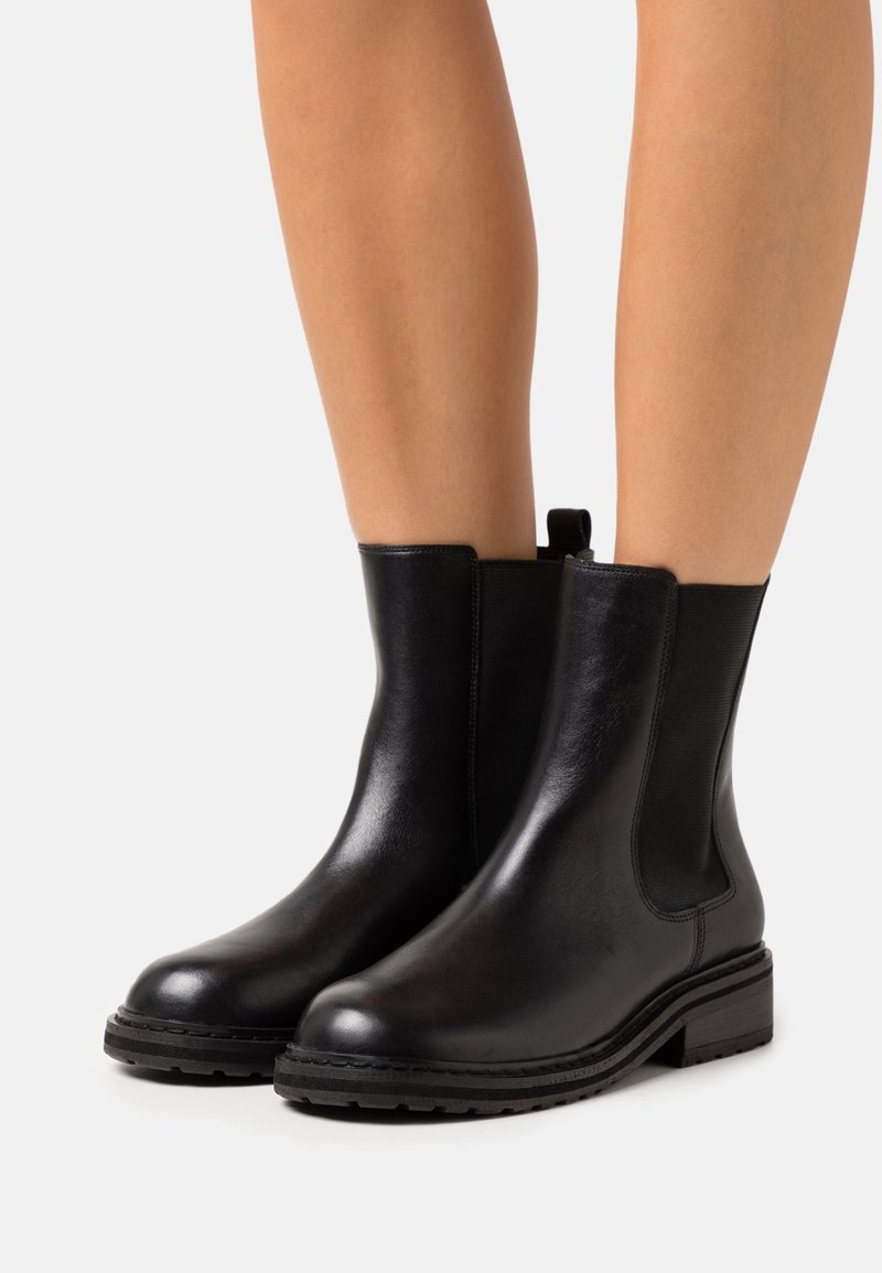 Bottes basses en cuir noir avec panneaux latéraux élastiques, bout rond et petit talon. Texture lisse avec une languette à l'arrière pour un enfilage facile.