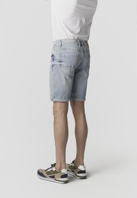 Férfi világoskék, visszahajtott farmer rövidnadrágban, hátsó zsebekkel, bézs, fekete és fehér sportcipőben, egyszerű háttér előtt állva.