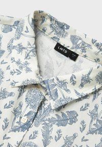 Camisa de manga corta con fondo crema y patrones de flores y hojas en azul, con cuello abotonado y una etiqueta textil que dice "LMTD."