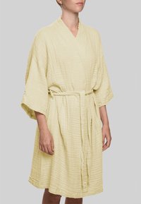 Šviesiai geltona chalatė iš tekstūruotos, raukšlėtos medžiagos. Turi plačias rankoves, juostelę ties juosmeniu ir siekia iki vidurio šlaunies. Paprastas dizainas.