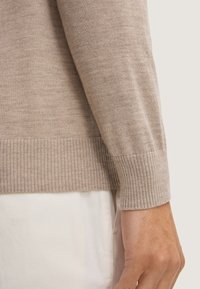 Beige gestrickter Pullover mit gerippten Bündchen und Saum, gefertigt aus einem weichen, strukturierten Stoff, kombiniert mit einer weißen Schicht darunter.