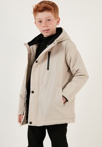 Beige, vadderad jacka med huva, svarta accenter, framfickor och en zip-försedd bröstficka. Mjuk inre foder; knapp- och dragkedjestängning.
