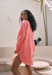 Rožinės spalvos siuvinėta marškinėliai ilgomis rankovėmis, šiek tiek laisvo silueto, su gėlių raštais ir užapvalintu apatine dalimi, dėvimi asmens šviesioje aplinkoje.