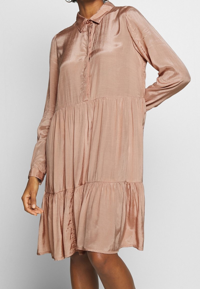 Donna che indossa un abito in satin a maniche lunghe, svasato e a strati di un delicato rosa chiaro, con colletto e bottoni frontali nascosti.