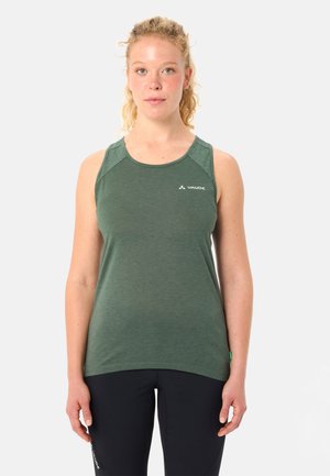 Donna con capelli ricci chiari, indossa una canotta sportiva verde senza maniche e pantaloni neri, in piedi contro uno sfondo chiaro uniforme, rivolta frontalmente.