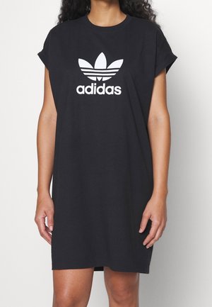 Kvinde iført en sort knælængde Adidas T-shirt-kjole med opkrullede ærmer og hvidt Adidas trefoil-logo på brystet.