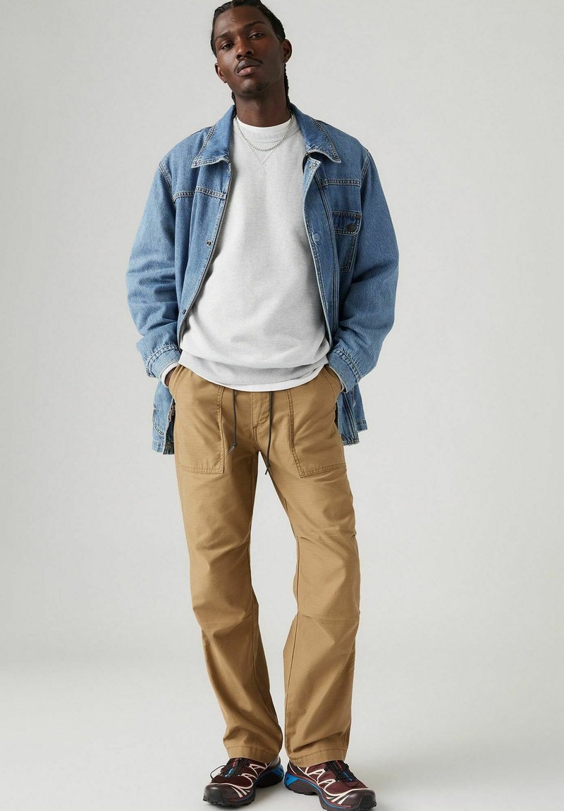 Chaqueta de mezclilla, sudadera gris y pantalones cargo en color beige. El atuendo presenta un corte holgado, con bolsillos laterales y detalles de costura en contraste.