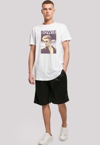 White cotton t-shirt featuring a graphic print of a man with sunglasses, text reading "BOWIE." Paired with black shorts and white sneakers.